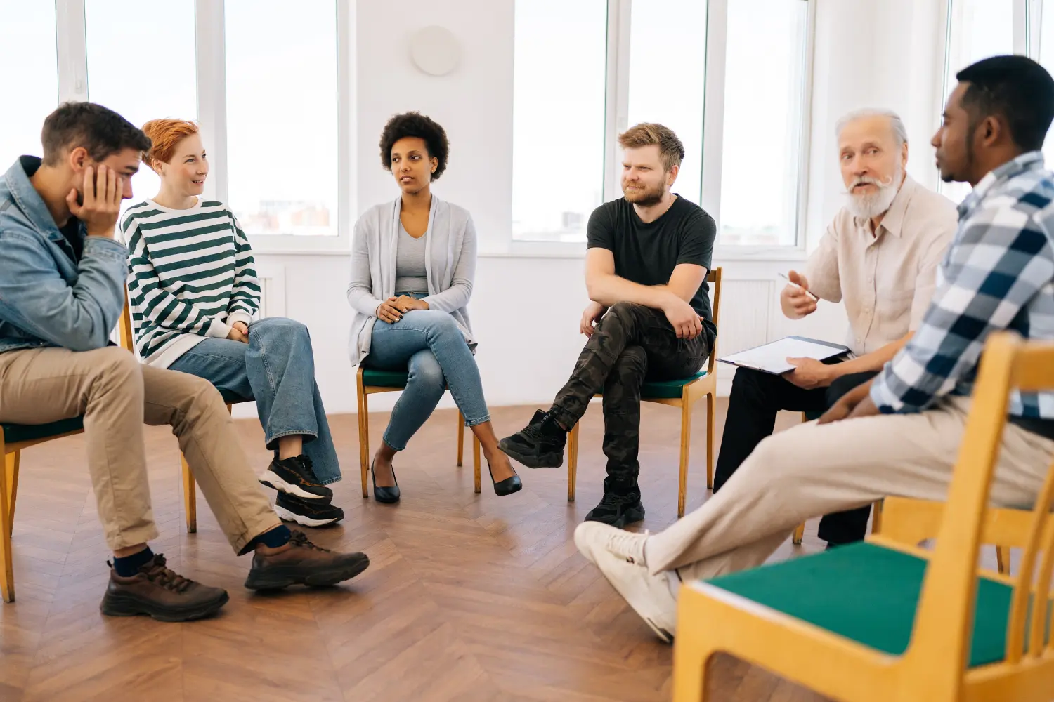 Weitwinkelaufnahme eines reifen männlichen Psychotherapeuten, der ein Therapietreffen mit süchtigen Menschen unterschiedlichen Alters leitet, die während des Therapietreffens im Kreis sitzen.